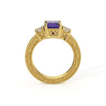 An ornate yellow gold ring featuring a rectangular velvet tanzanite center stone, flanked by small clear stones and tiny red accent gems, set on a highly detailed beaded filigree band.