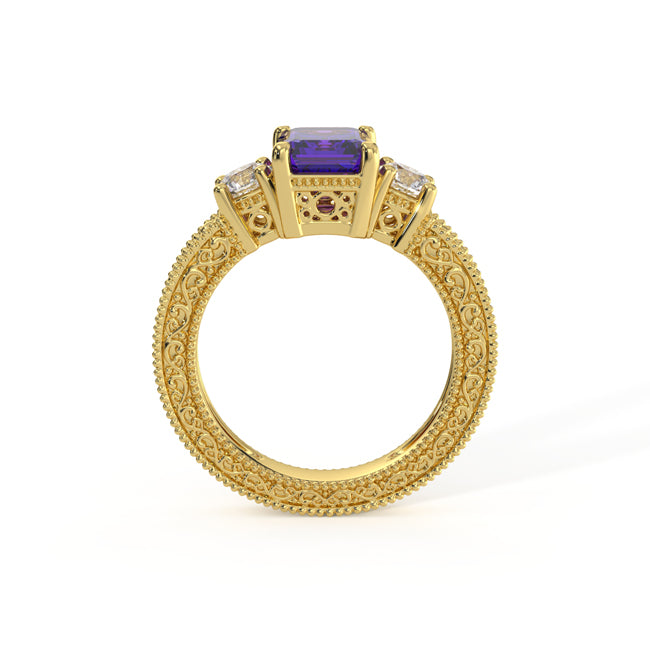 An ornate yellow gold ring featuring a rectangular velvet tanzanite center stone, flanked by small clear stones and tiny red accent gems, set on a highly detailed beaded filigree band.