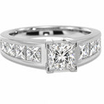 A white-toned diamond ring featuring a large princess-cut center stone with channel-set diamonds on either side, shown on a clean white background.