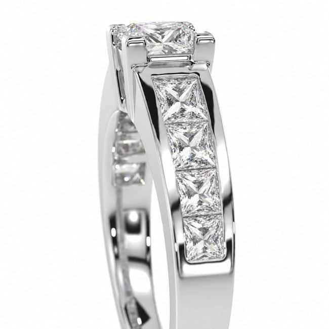 A close-up studio shot of a white gold or platinum ring featuring a princess-cut diamond center stone and a channel setting of smaller princess-cut diamonds along the band, set against a plain white background.
