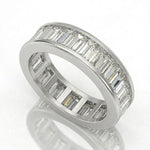 A white-colored eternity ring featuring a channel setting of baguette-cut clear stones on one side and round-cut stones on the other, displayed on a reflective white surface.